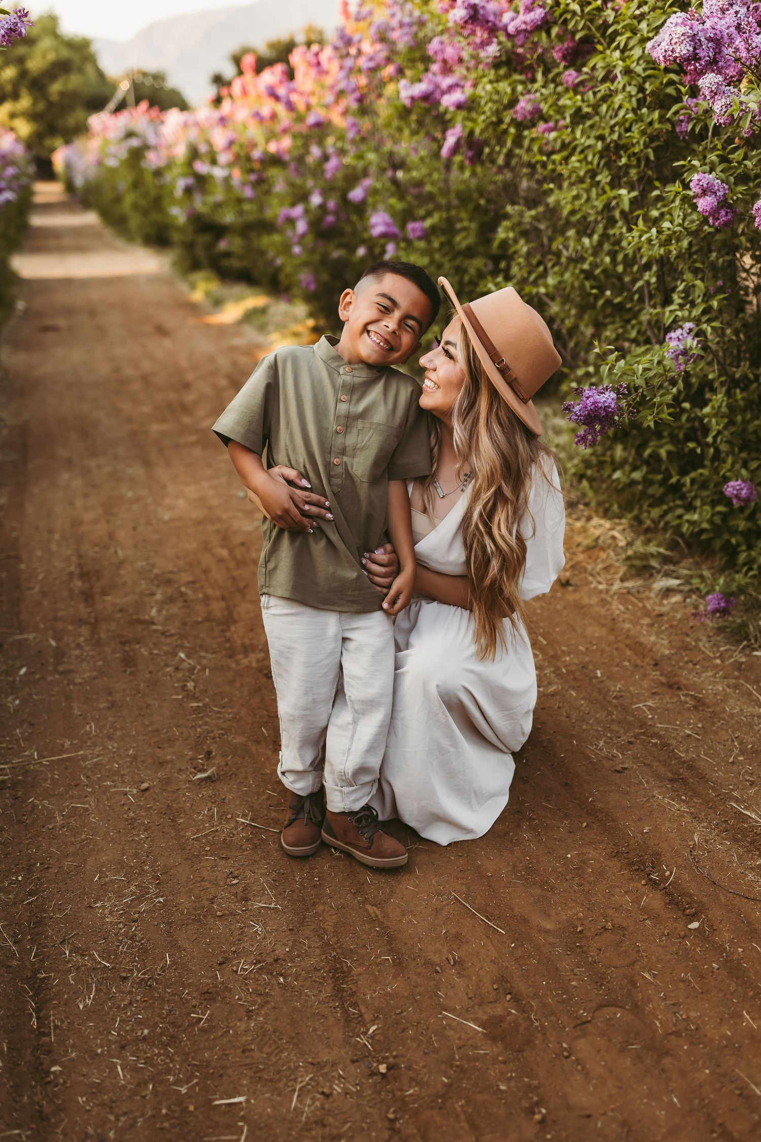 A mom and son embraces and laughs together along a dirt path lined with blooming purple lilac bushes.