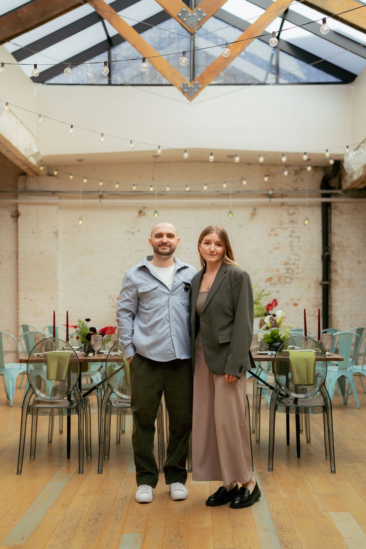 A rustic event space with exposed wooden beams, string lights, and mint green chairs arranged at tables beneath a glass ceiling.