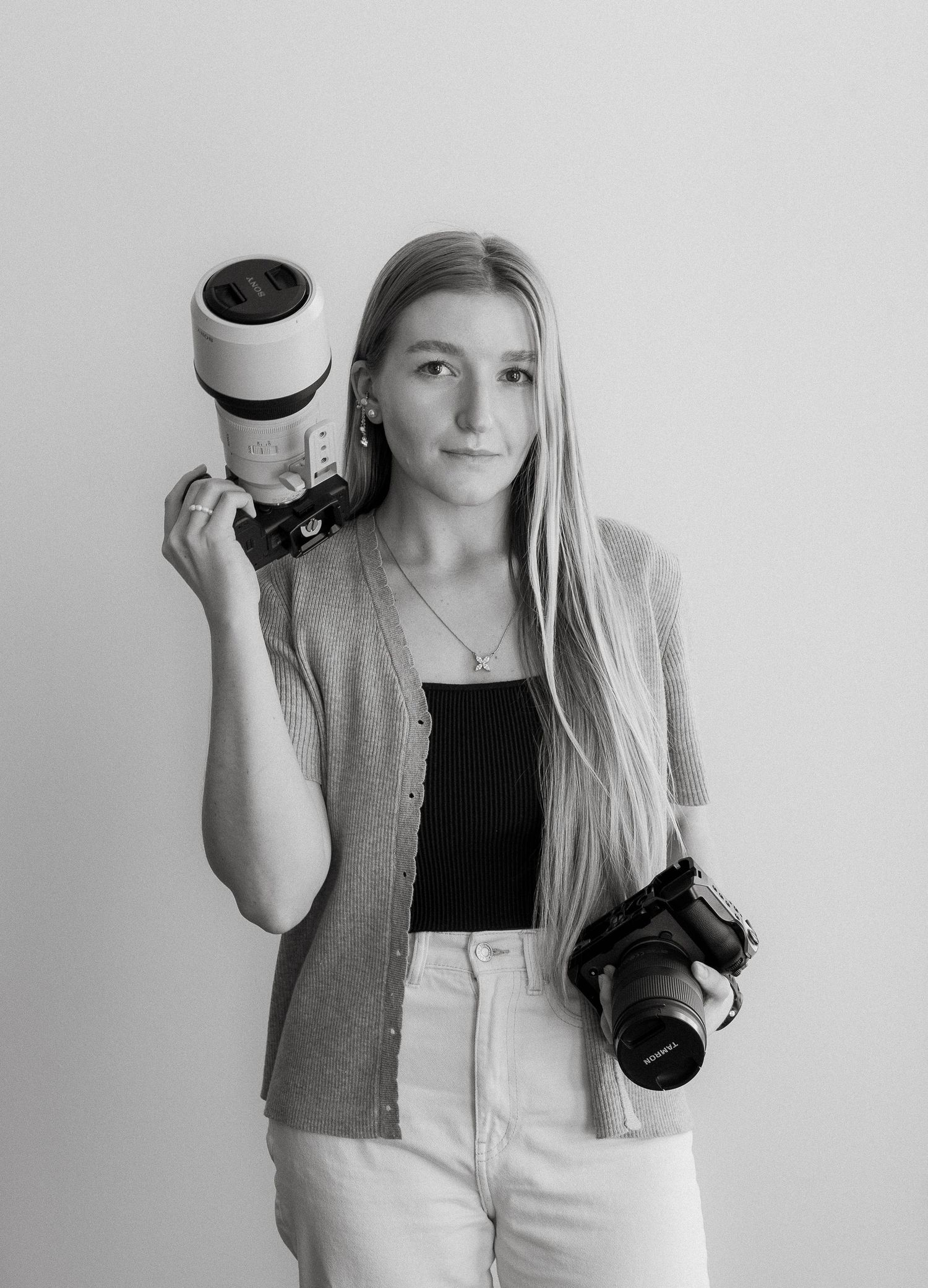 Professional photographer holding two DSLR cameras with long lenses while wearing a gray cardigan and white pants.