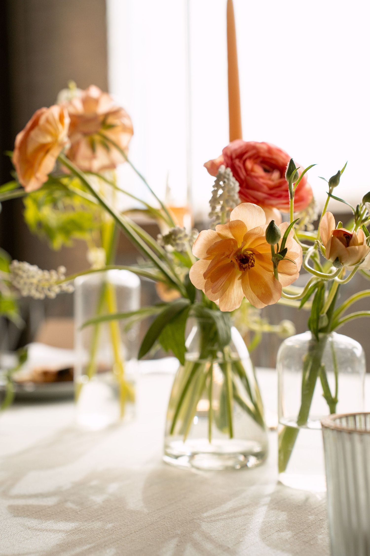 Fresh peach and coral flowers arranged in clear glass vases with delicate white blooms against a bright window backdrop.