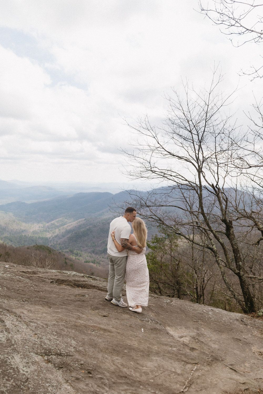 Two people share an intimate moment while overlooking a scenic mountain vista from a rocky overlook on a cloudy day.