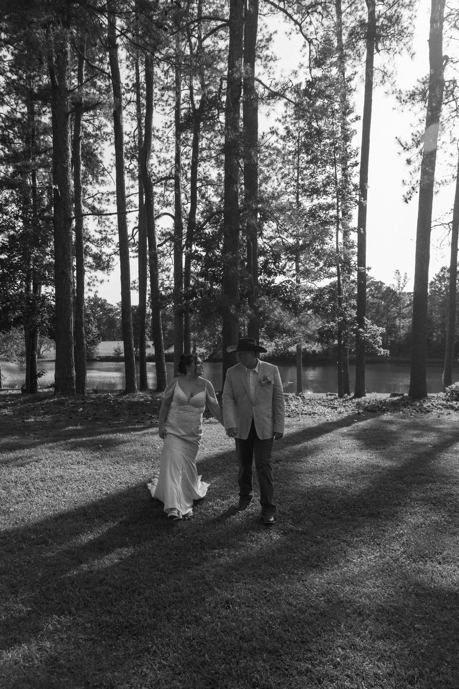 Black and white wedding photo of couple standing together on a wooded path with tall pine trees and sunlight filtering through.