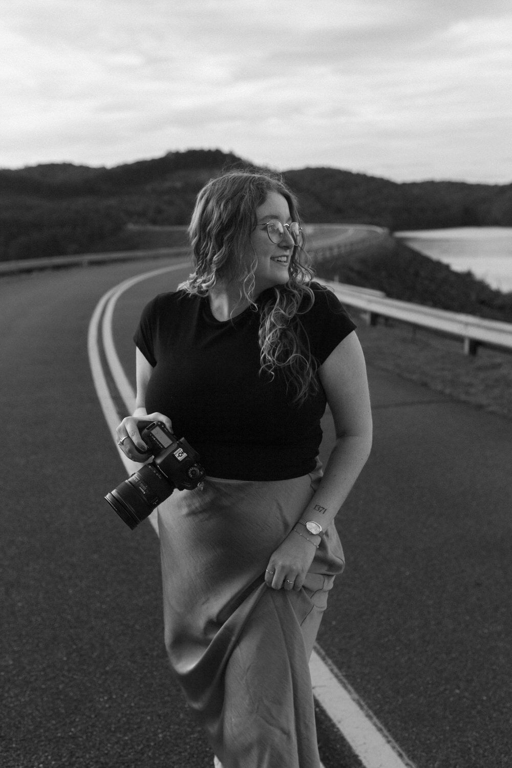 A joyful photographer, Sydney, in black top holds a camera while standing on a winding mountain road during sunset.