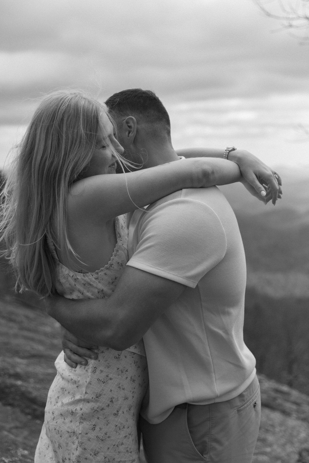 Romantic black and white photograph of a couple embracing while standing on a hillside against a cloudy sky.