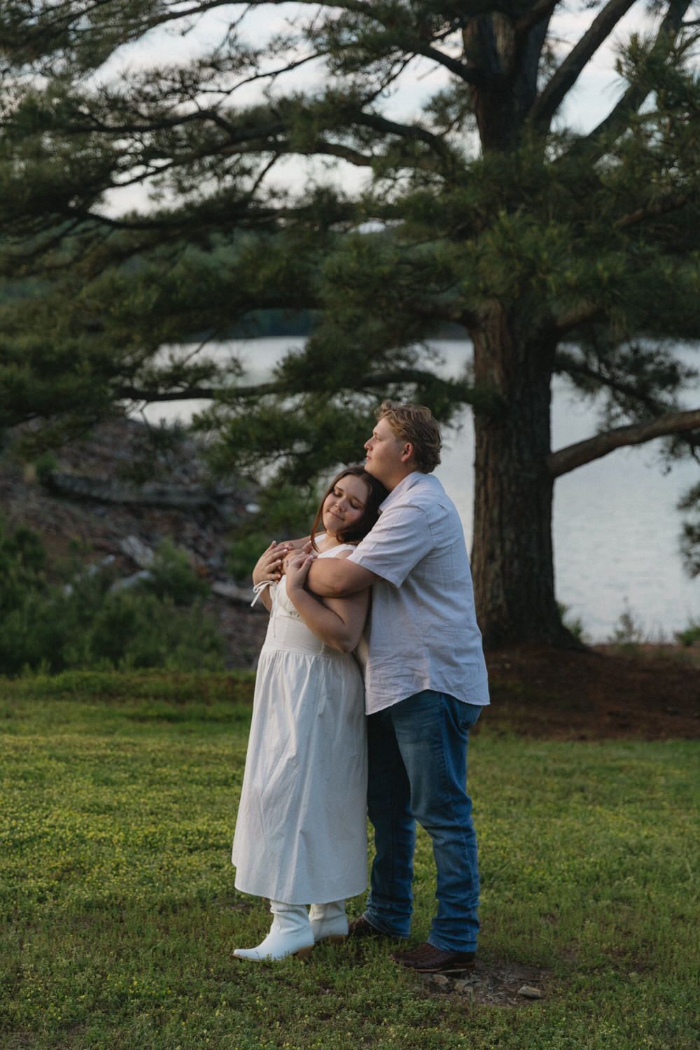 A couple embraces lovingly under a tall pine tree on a grassy hillside overlooking a lake at dusk in the appalachian mountains