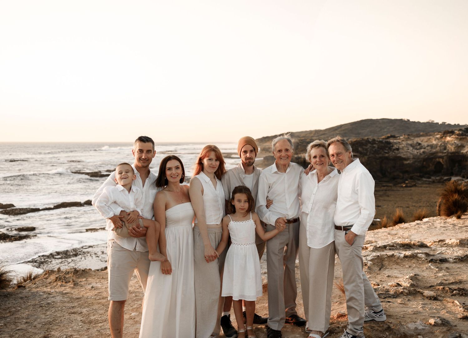 a large family standing on top of a cliff in front of the ocean looking towards the camera smiling and wearing neutral coloured clothing