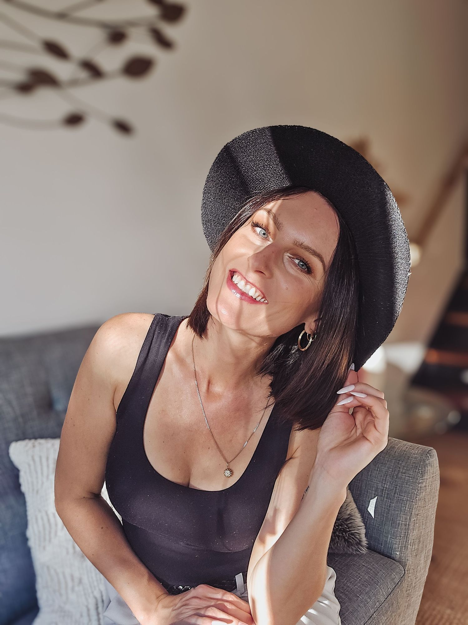 a smiling woman with blue eyes sitting down wearing a black hat whilst holding the brim