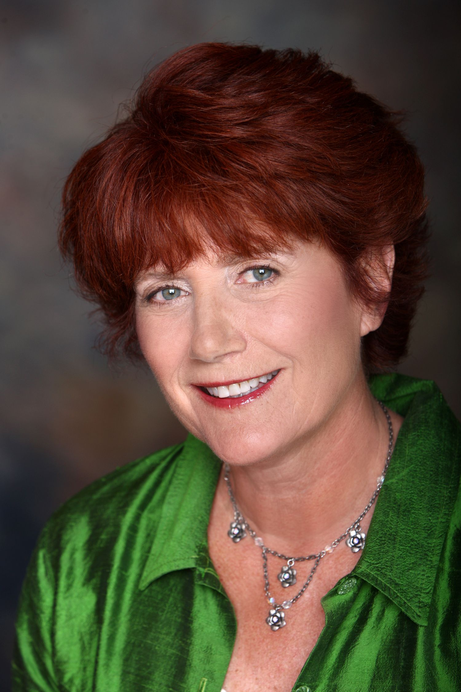 Professional headshot of a woman with short red hair wearing a bright green top and statement necklace.