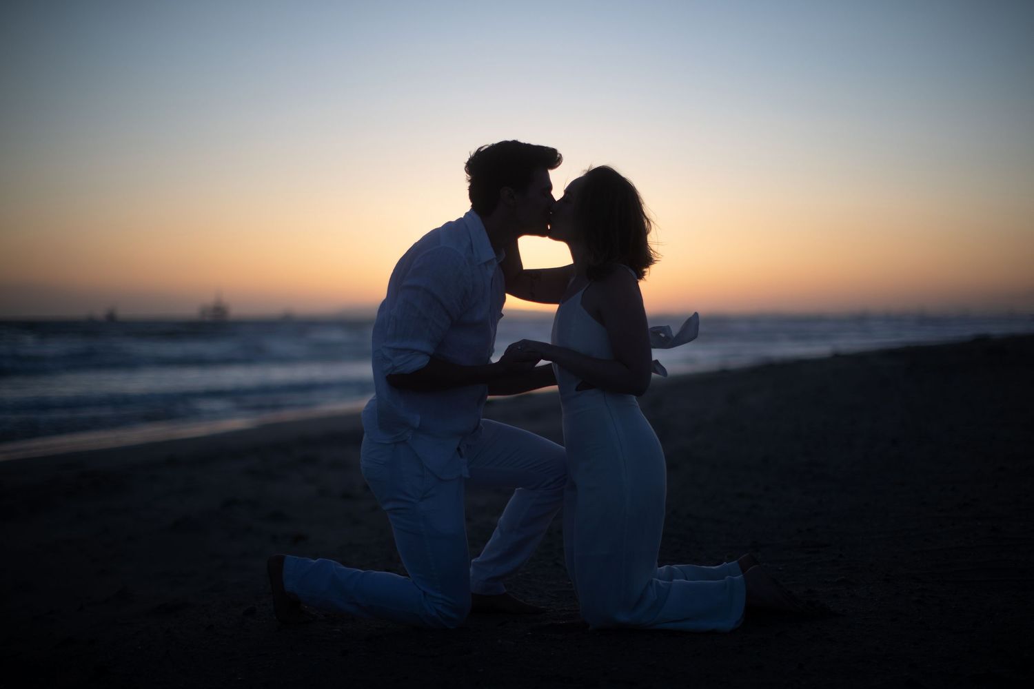 Two silhouetted figures share a romantic moment on a beach at sunset as ocean waves roll in the background.