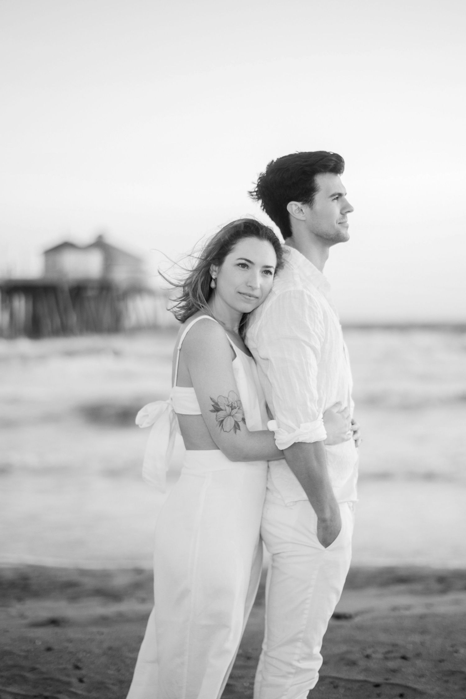 Couple in white outfits pose for romantic black and white photo on beach with pier in background.