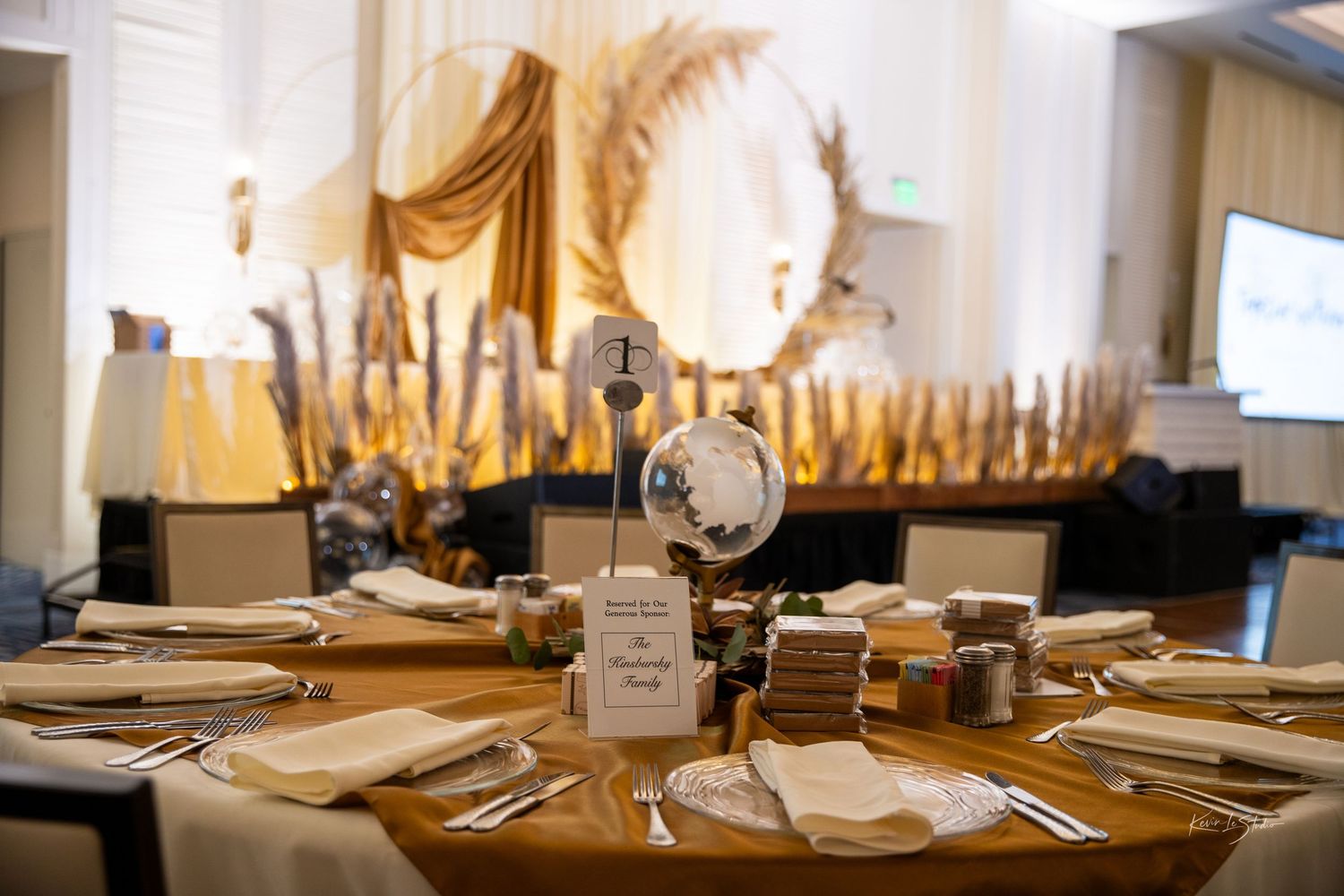 Elegant golden wedding reception table settings with white linens and warm ambient lighting in a banquet hall.