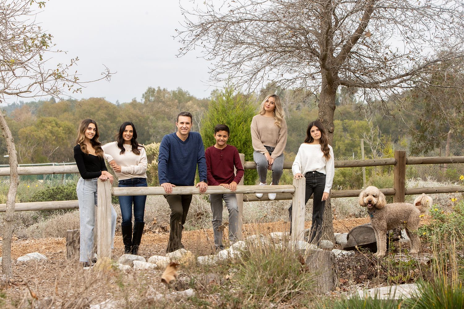 Family casually posed on wooden fence with dog in outdoor nature setting.