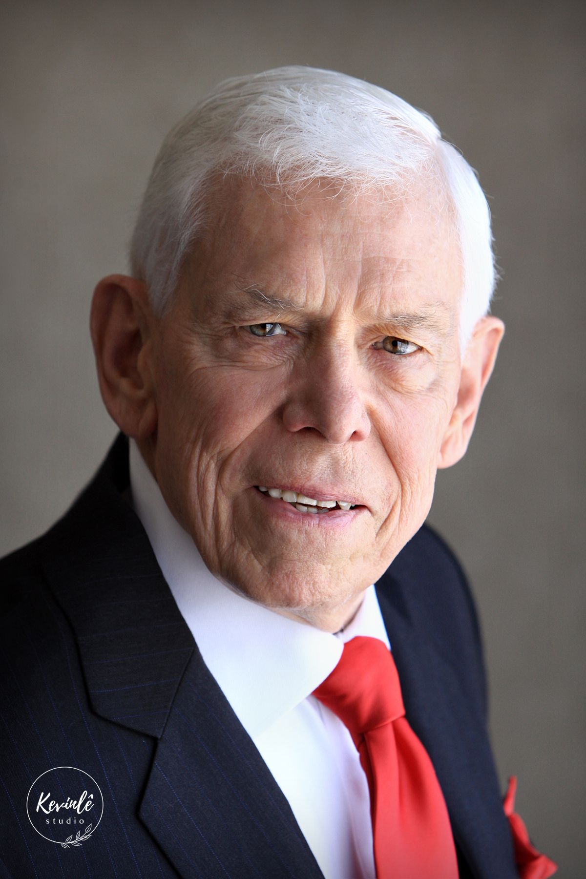 Professional portrait of an older gentleman with white hair wearing a dark suit and red tie.