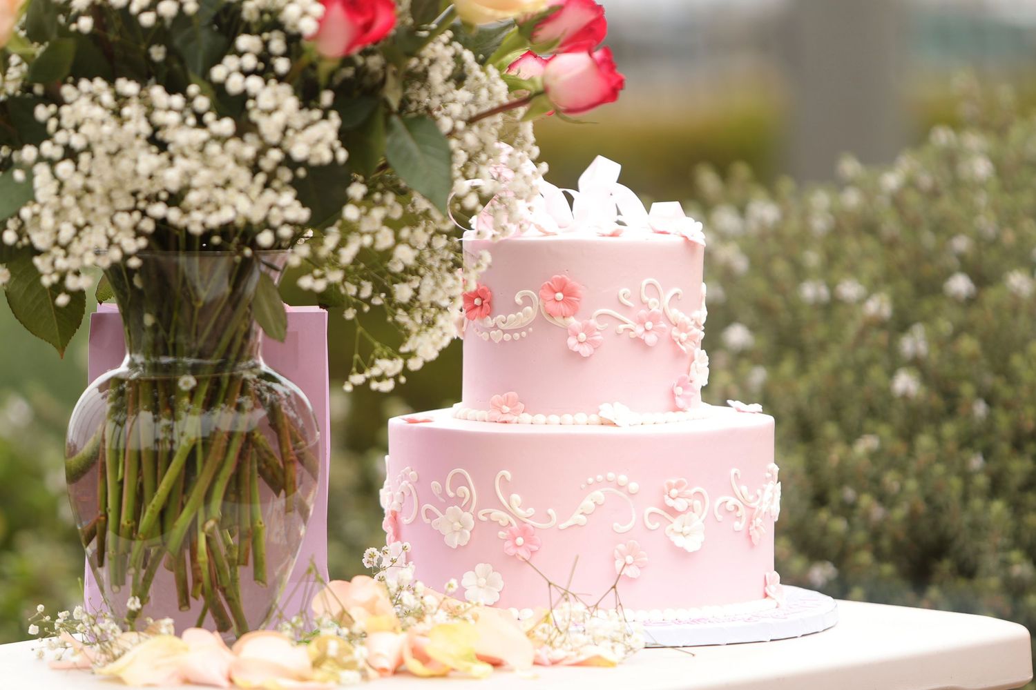 A delicate pink two-tiered wedding cake with floral decorations sits next to a vase of flowers in an outdoor setting.