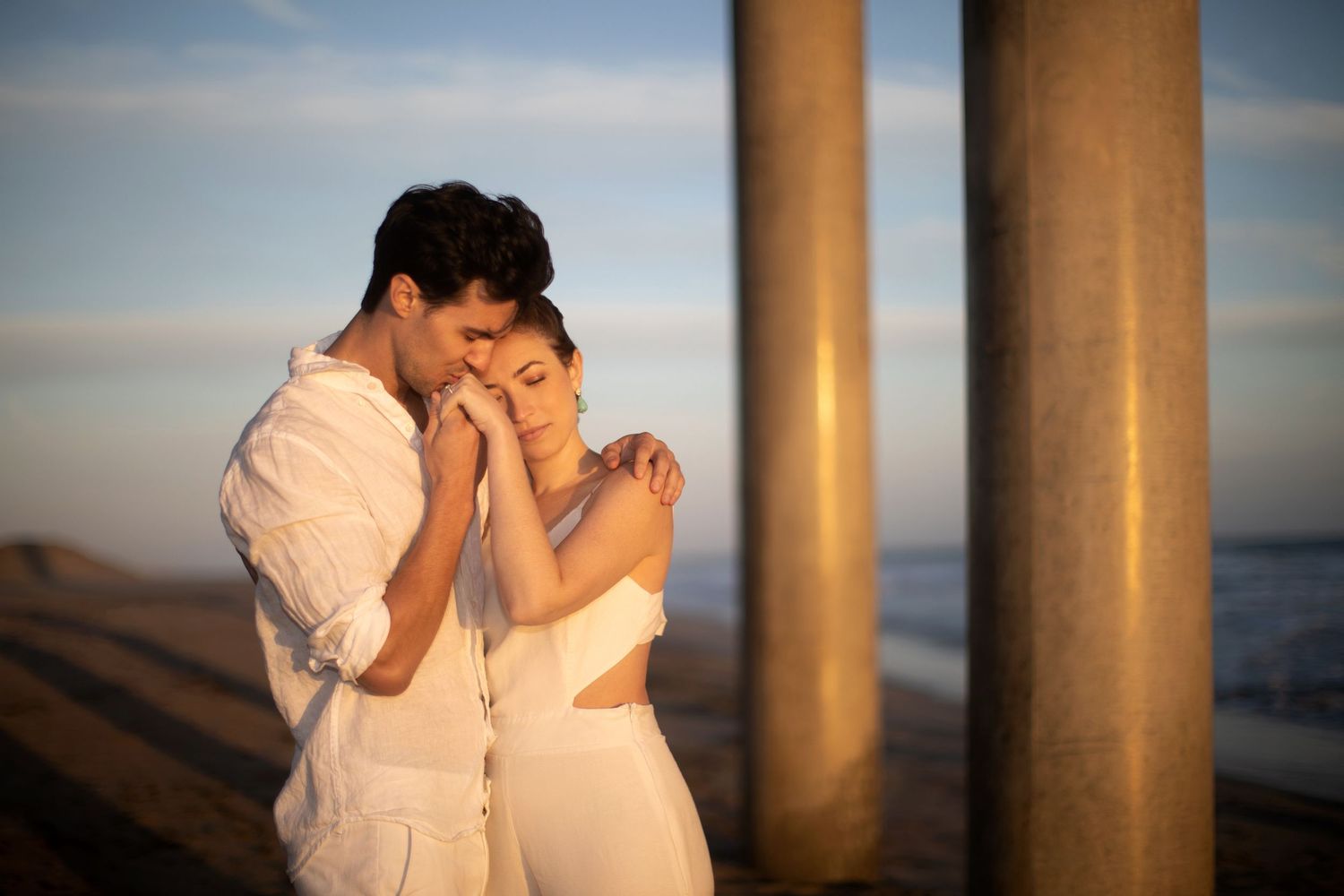 Couple in white outfits sharing tender moment between pier pillars at golden hour.
