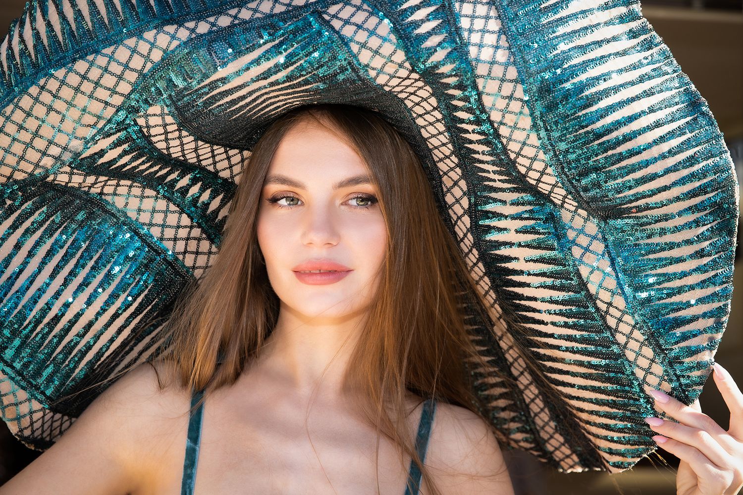 Model wearing a large metallic turquoise hat in natural lighting.