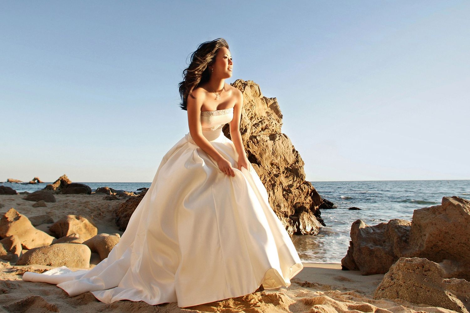Bride in flowing white gown poses dramatically on rocky beach at sunset.