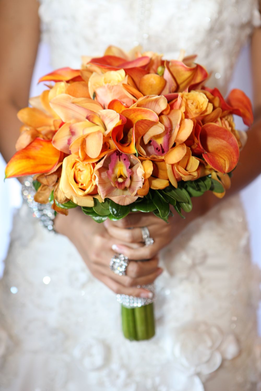 A vibrant orange bridal bouquet with roses and orchids wrapped in silver ribbon against a white lace dress.