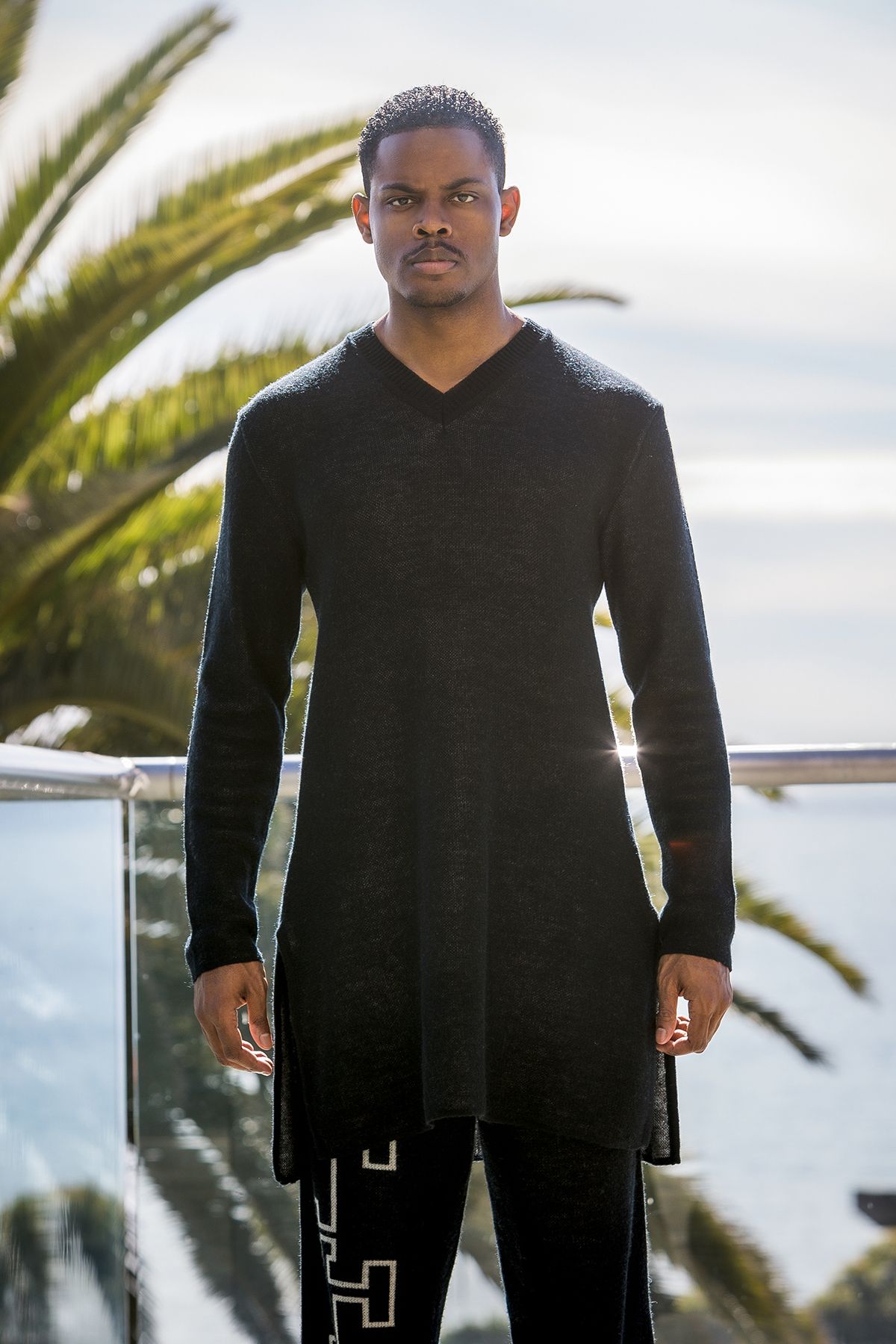 Model wearing black v-neck sweater stands outdoors near palm trees.