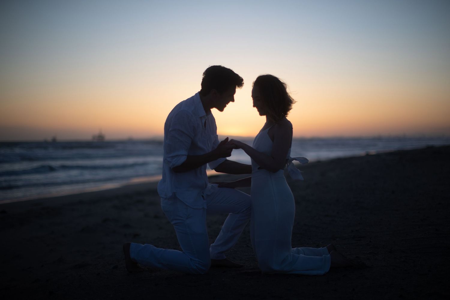 Couple kneeling together on beach during sunset engagement moment.