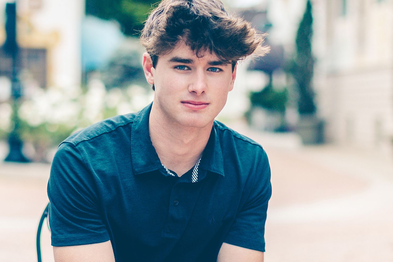 A person wearing a navy blue t-shirt poses casually in an outdoor setting with soft lighting and a blurred urban background.