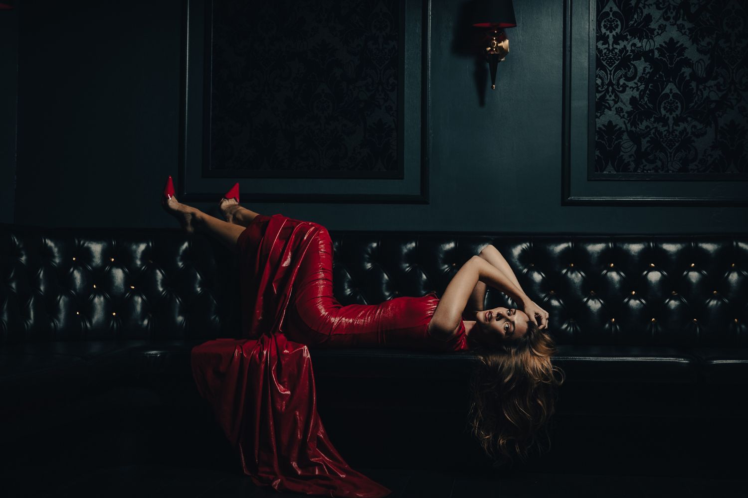 Model in flowing red dress reclining dramatically on dark leather couch.