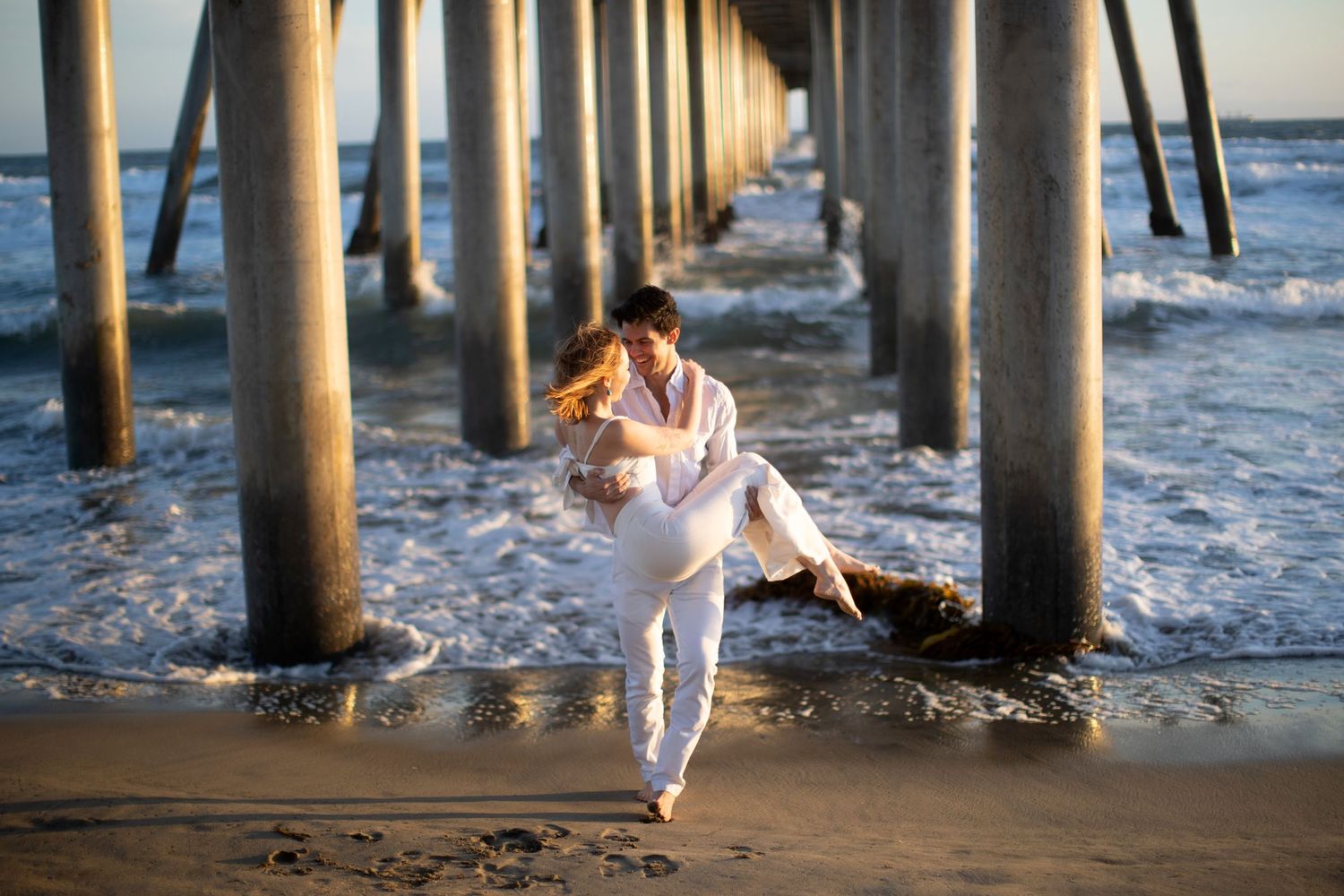 Boyfriend in white carrying partner under beach pier as waves crash nearby.