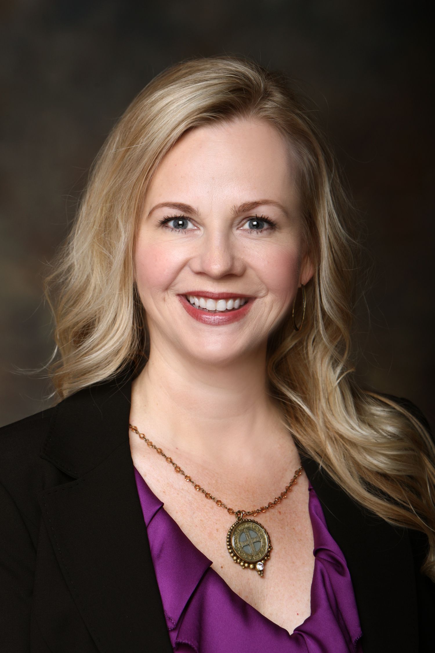 Professional headshot of person in purple blouse and black blazer with pendant necklace.