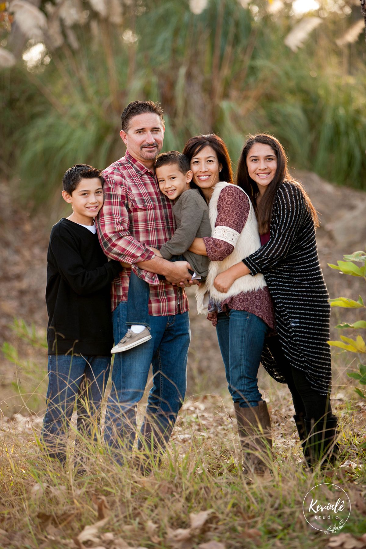 A family poses together outdoors in casual attire with fall foliage in the background.