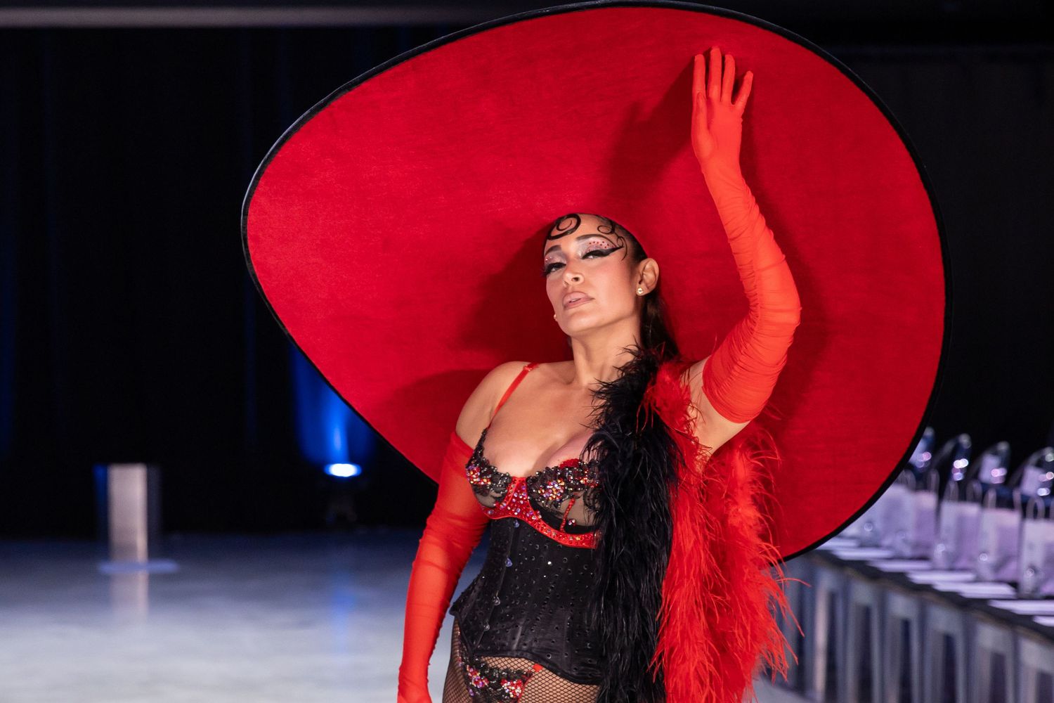 A model wearing a dramatic red hat and black lace ensemble poses on a runway during a fashion show.