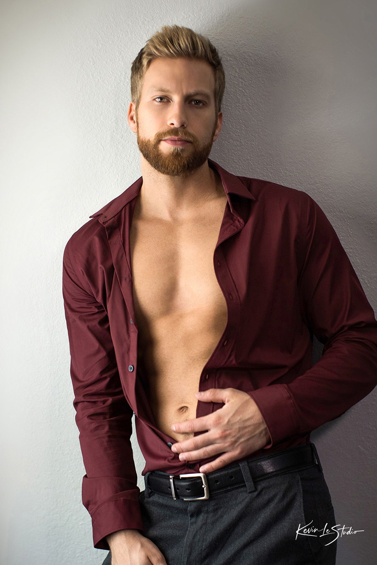 Fashion portrait of muscular model in burgundy shirt against white wall.