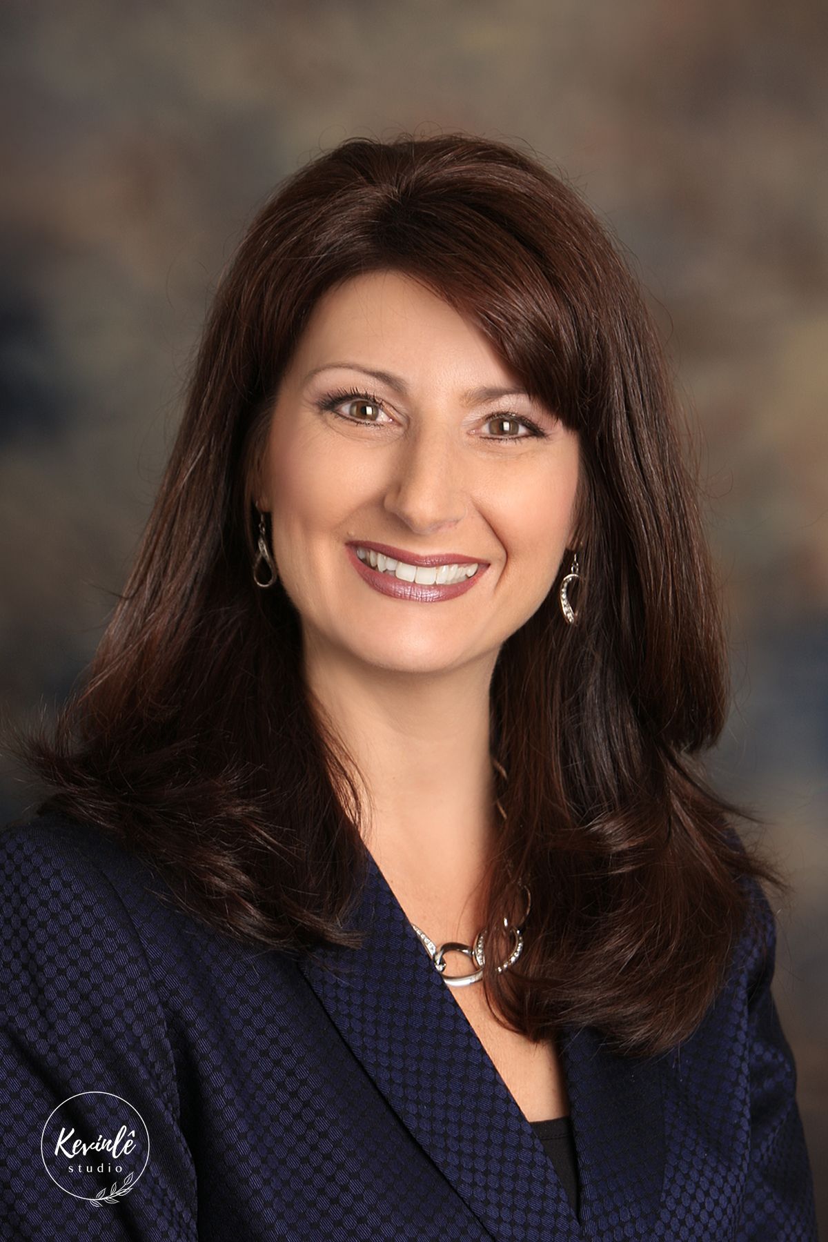 Professional portrait of a woman with long dark hair wearing a navy blazer and drop earrings.