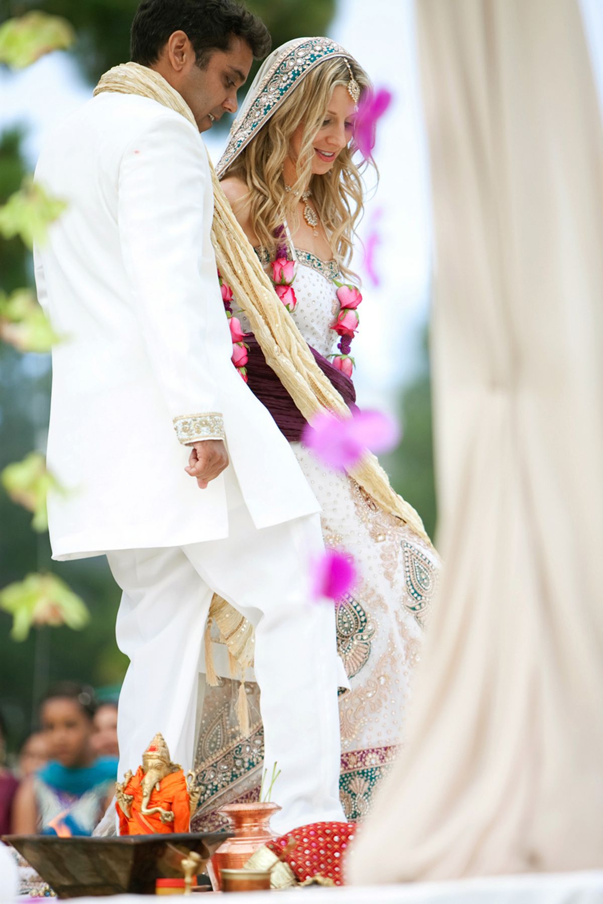 Couple in white attire participates in cultural wedding ceremony with colorful floral accents.