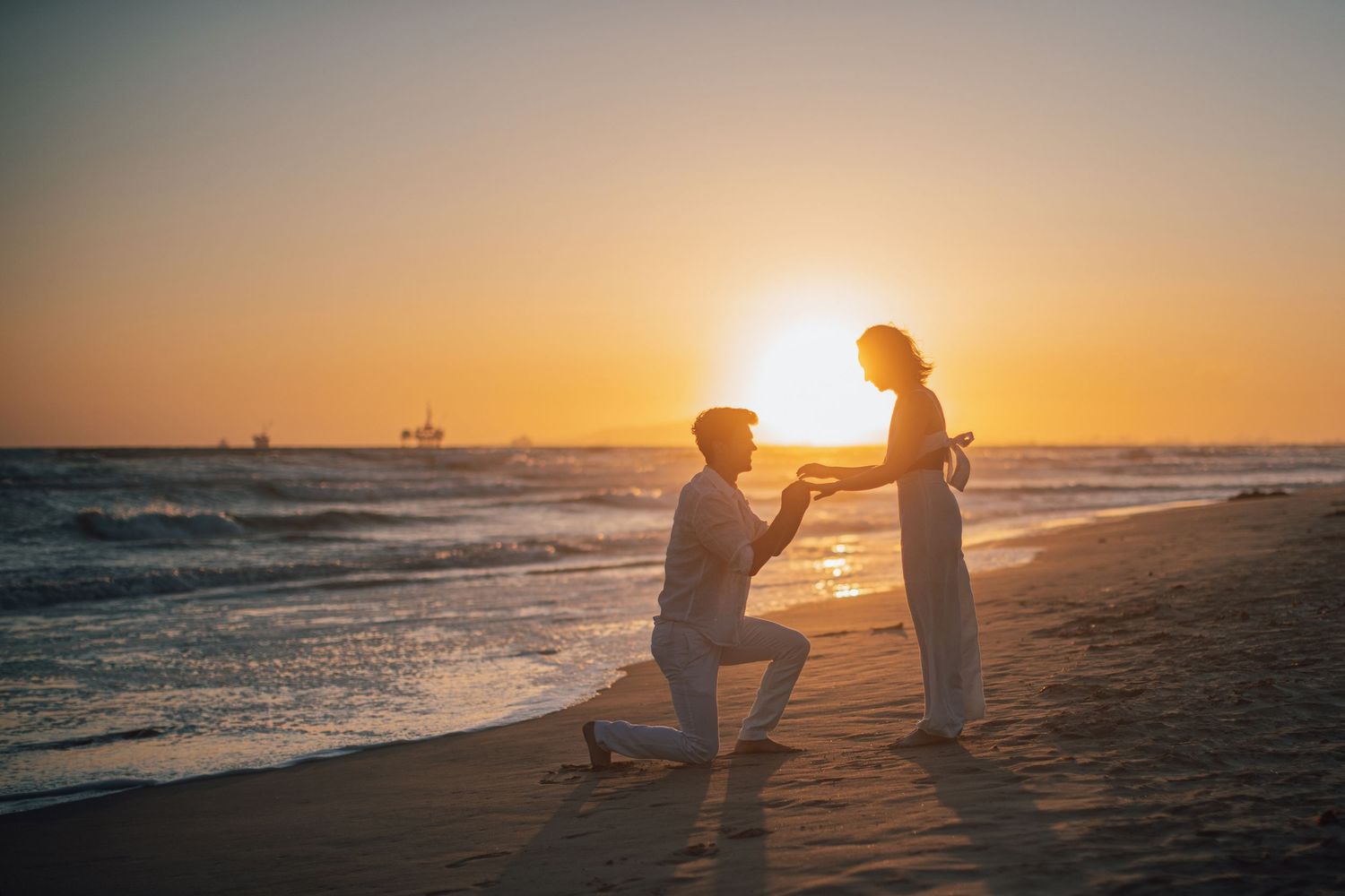 Silhouette of marriage proposal on beach at sunset with ships in background.