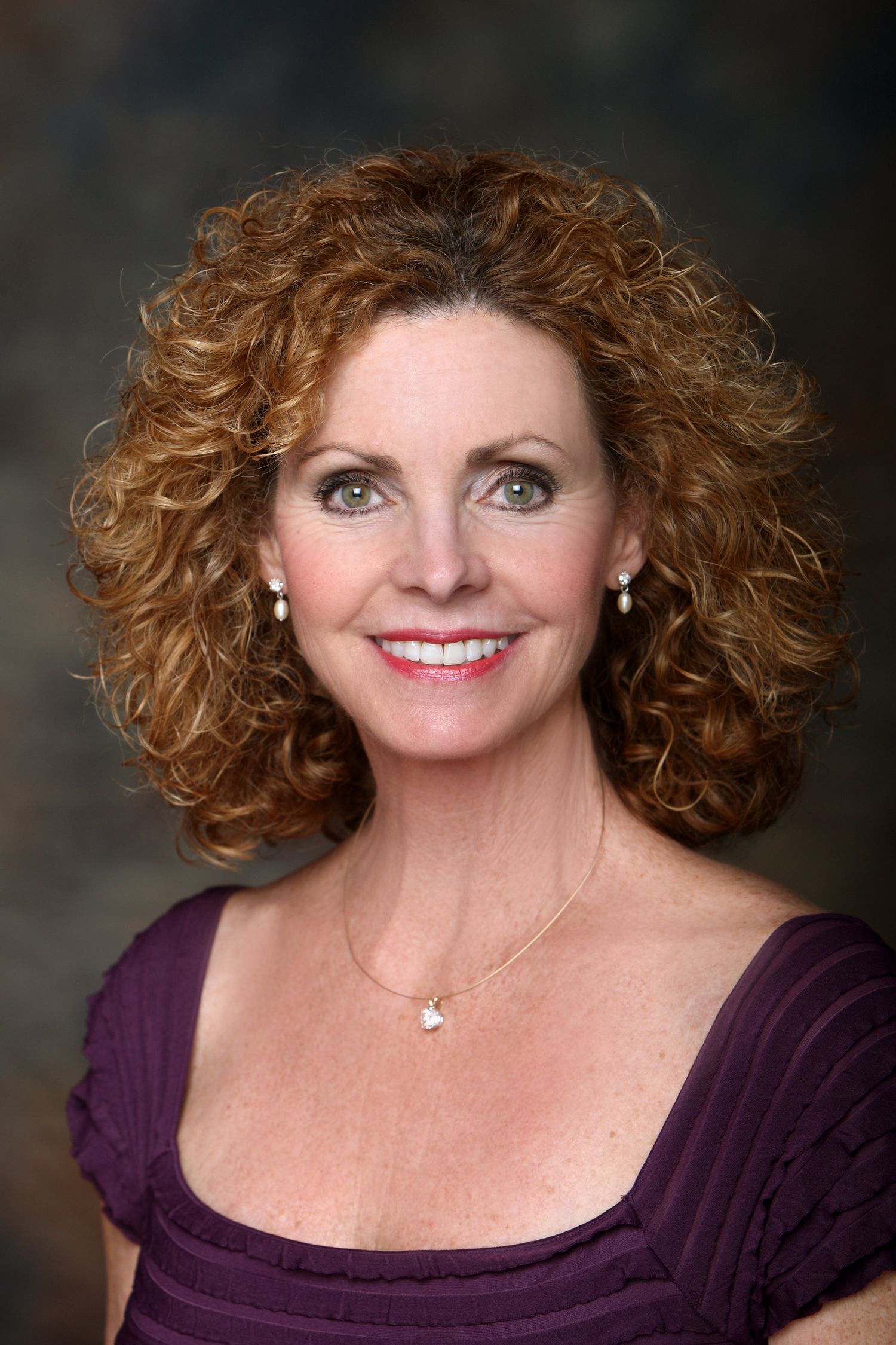 Professional headshot of a woman with curly honey-blonde hair wearing a purple top and delicate necklace.