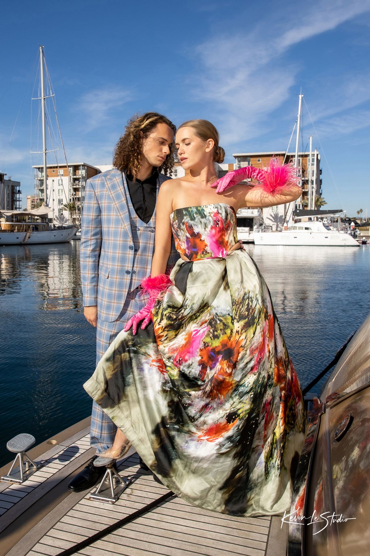 Two models pose affectionately on a wooden dock with sailboats and a marina in the background on a sunny day.