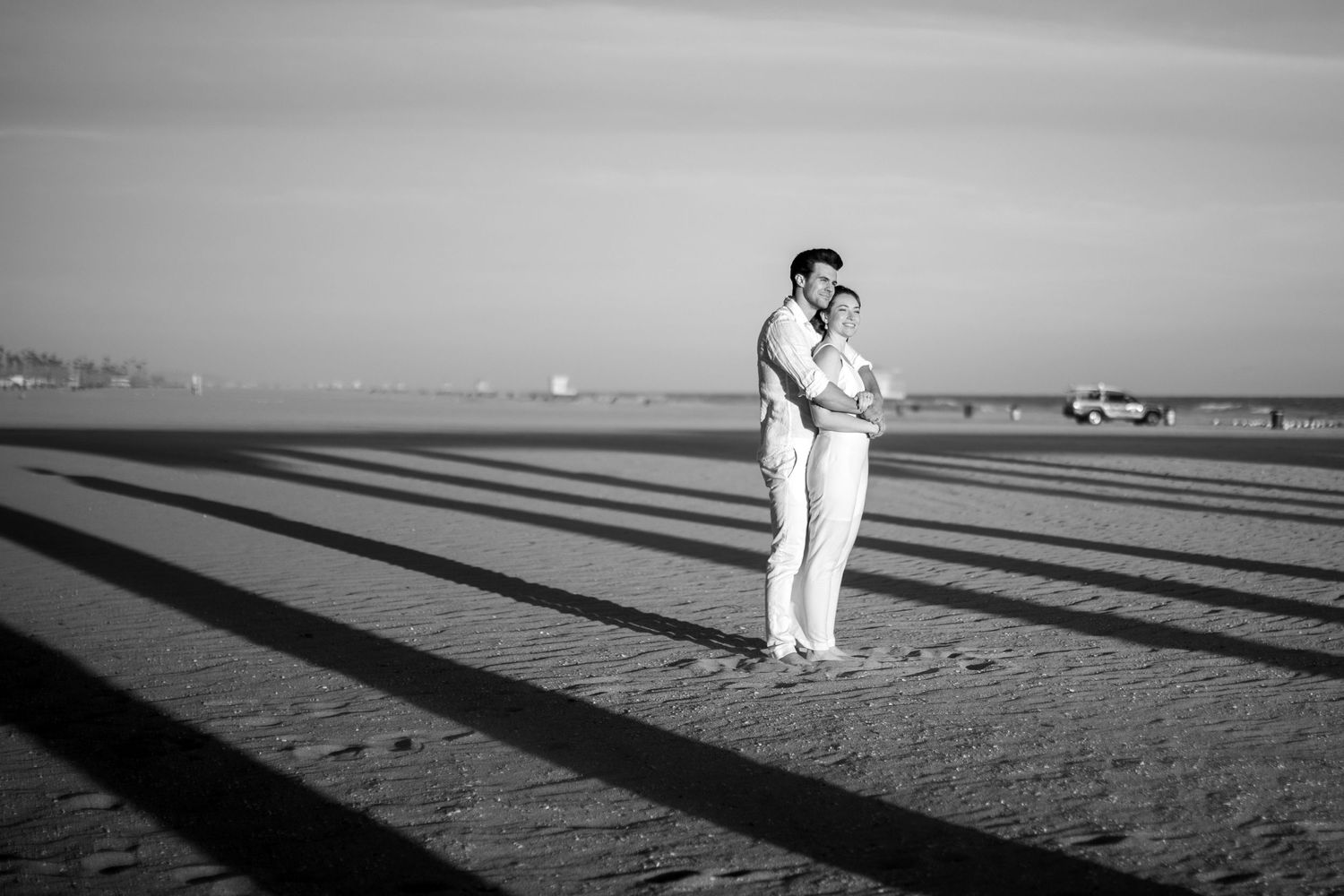 Black and white photo of couple embracing on beach with dramatic shadows from pier.