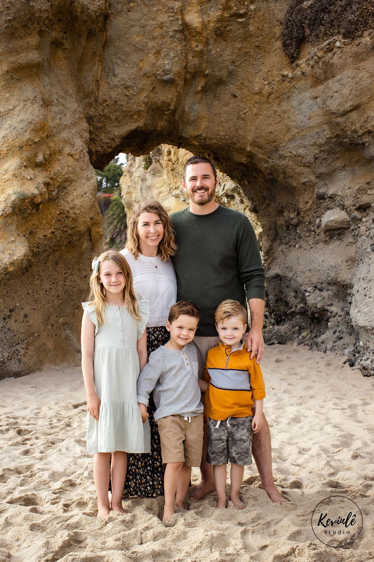 Happy family of five poses together under natural rock arch formation on sandy beach.