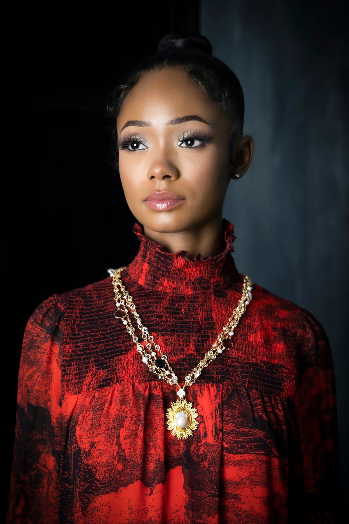 Portrait in red turtleneck sweater with ornate gold necklace against dark background.