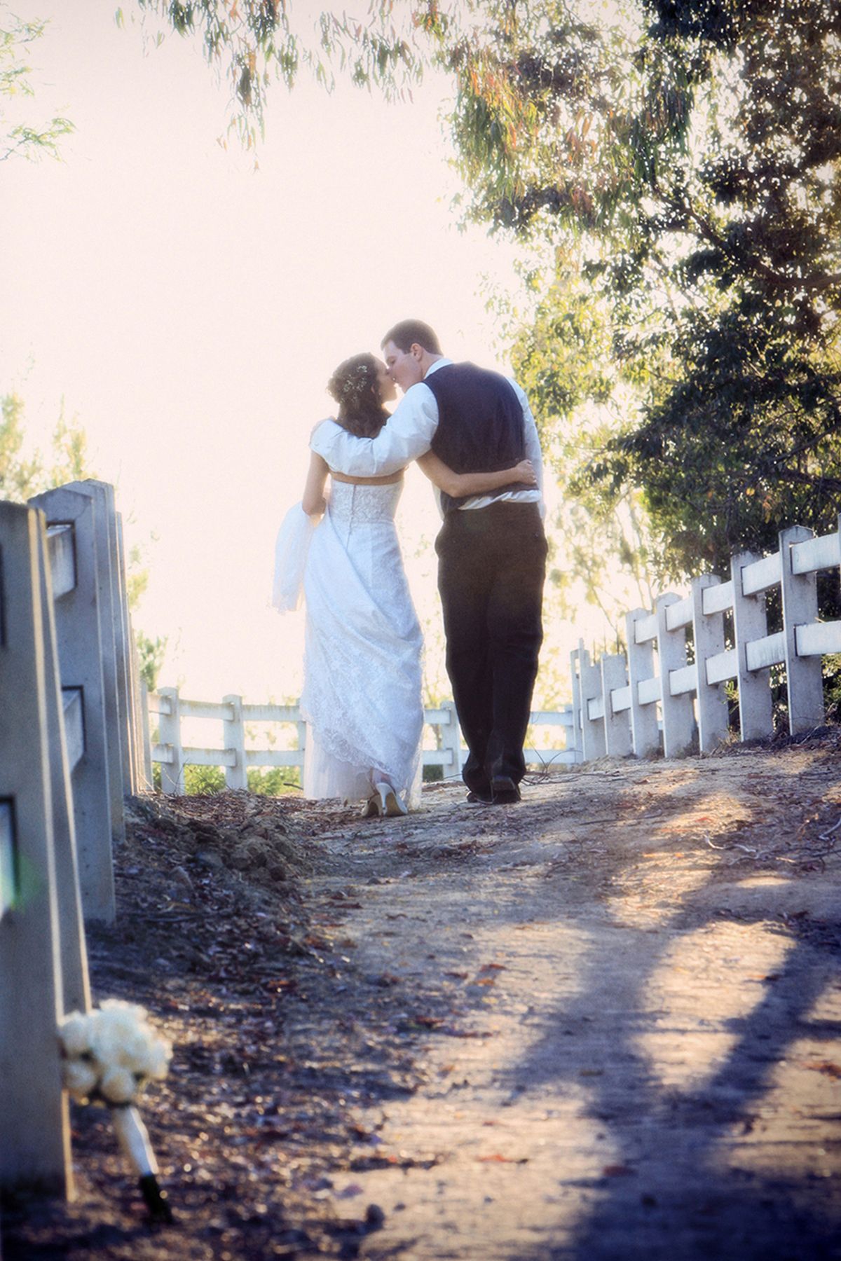 Couple walking along a sunlit path with white fence and trees in background.