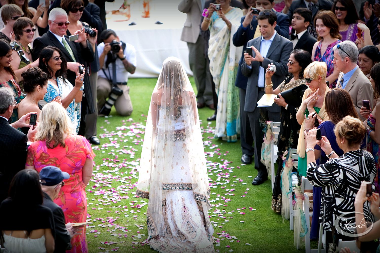 Wedding guests watching ceremony on lawn with flower petals scattered along aisle.
