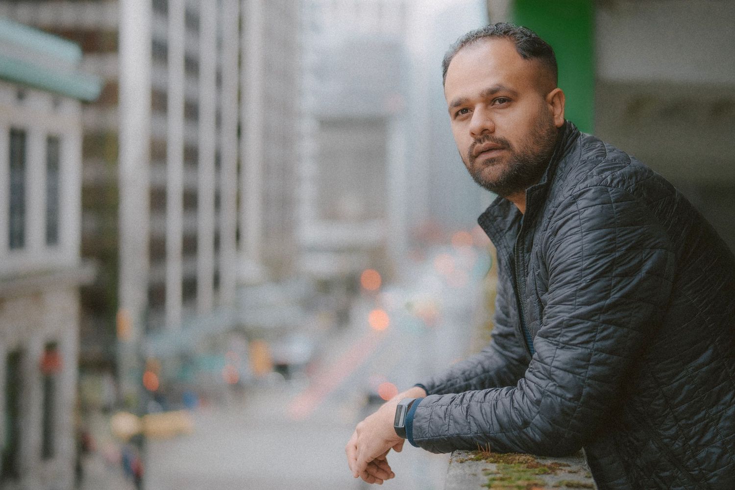 Person wearing a quilted black jacket leans against an industrial metal railing in an urban setting.