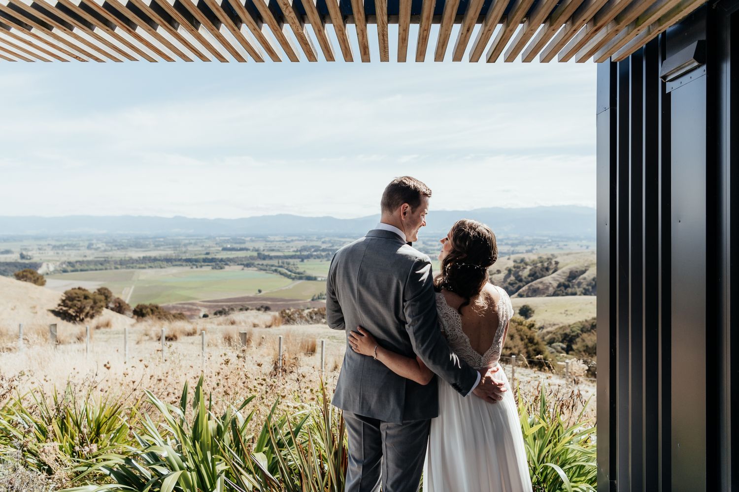 A couple in wedding attire embrace while admiring a scenic valley vista from a modern covered terrace at Whitimanuka by August and Vine