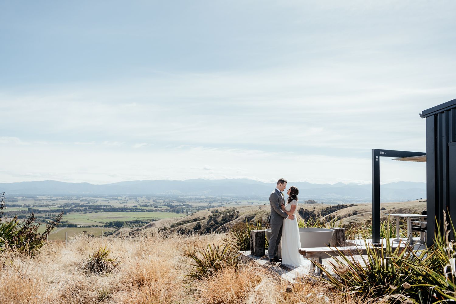 Couple stands at scenic vista at Whitimanuka Martinborough on wedding day overlooking vast rural landscape from deck by August and Vine