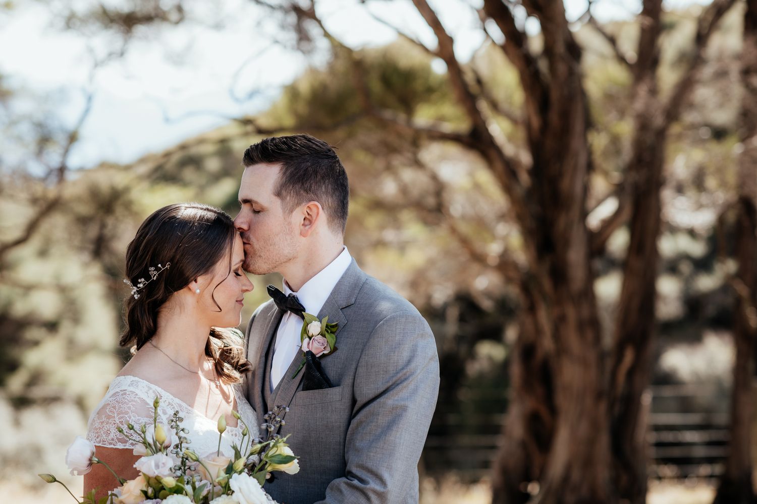 A romantic outdoor wedding moment between a couple in a gray suit and lace dress amid rustic natural scenery by August and Vine