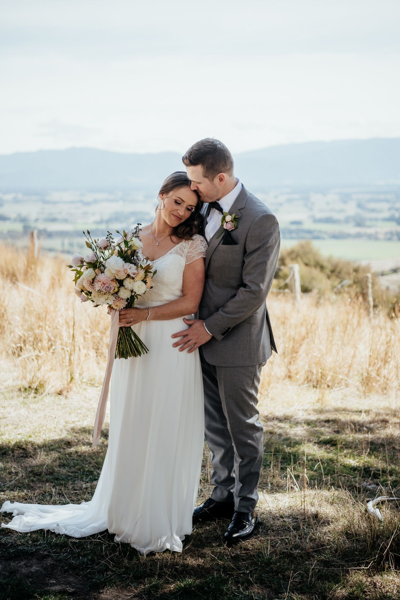 A newlywed couple embraces on a hilltop with mountain views at Whitimanuka, bride in white gown holding floral bouquet by August and Vine