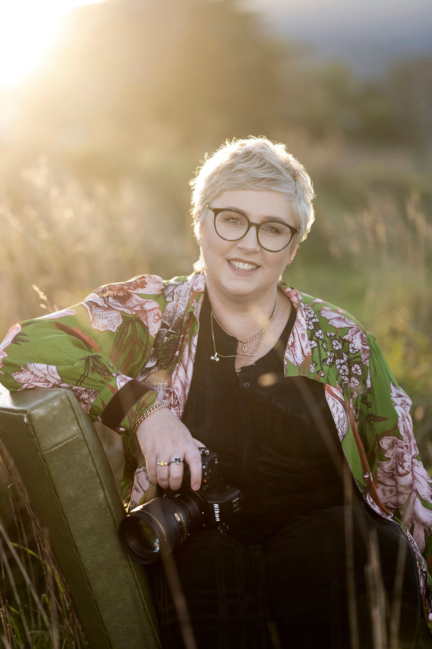 Professional photographer August and Vine with short blonde hair, glasses and floral jacket holding nikon camera at sunset in a field.