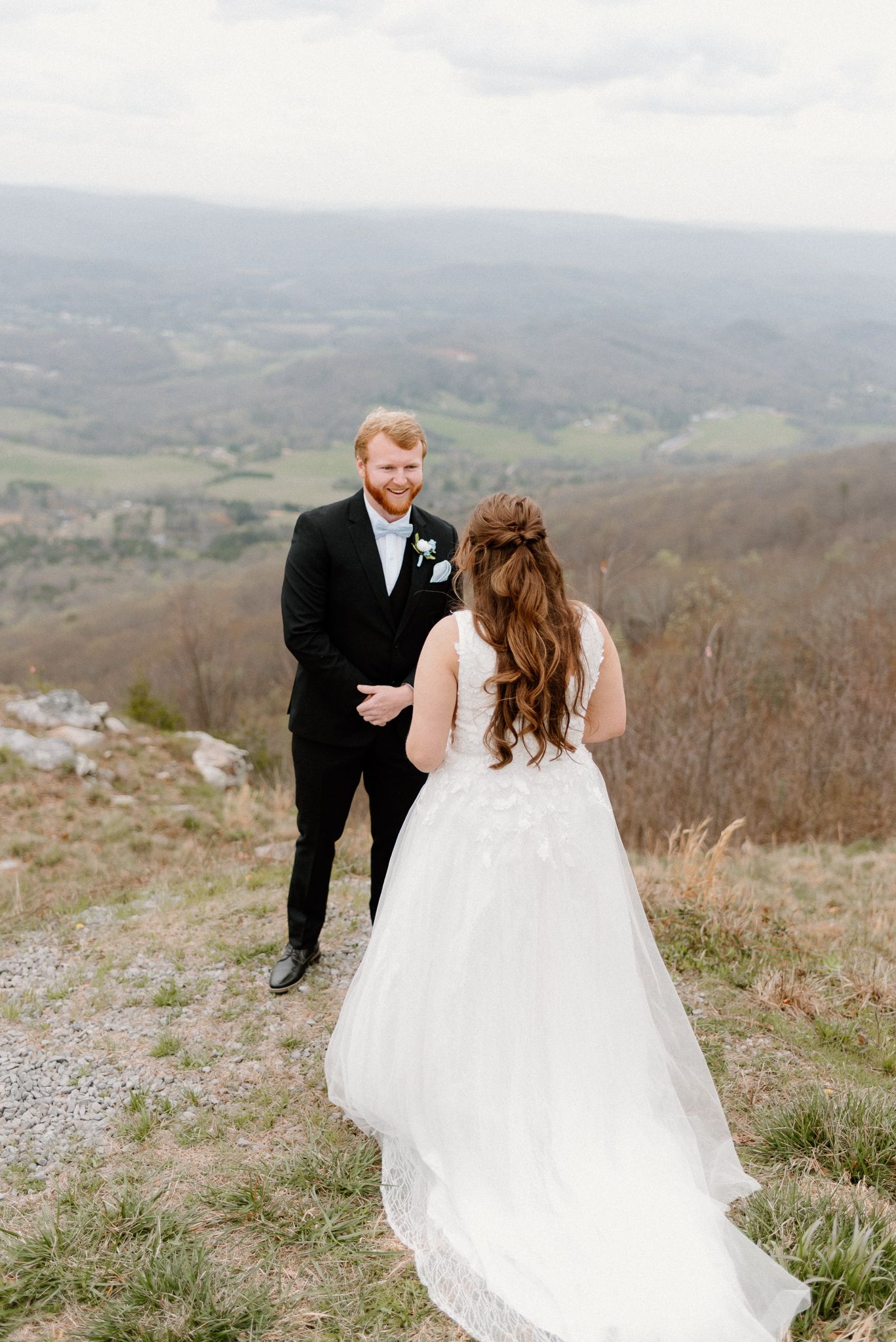 A couple shares a magical first look moment atop a mountain overlook with sweeping valley views in the distance.