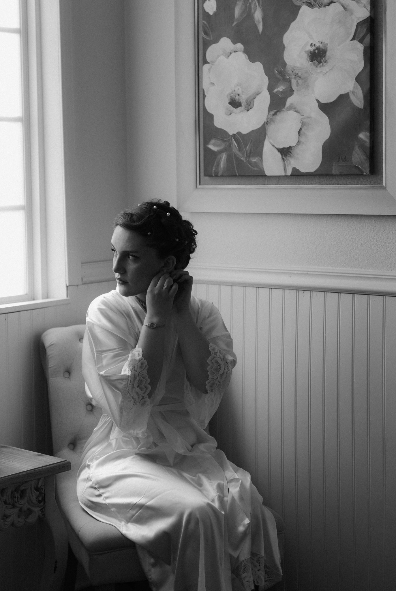 Black and white artistic photograph of someone in white garments seated by a window beneath floral artwork.
