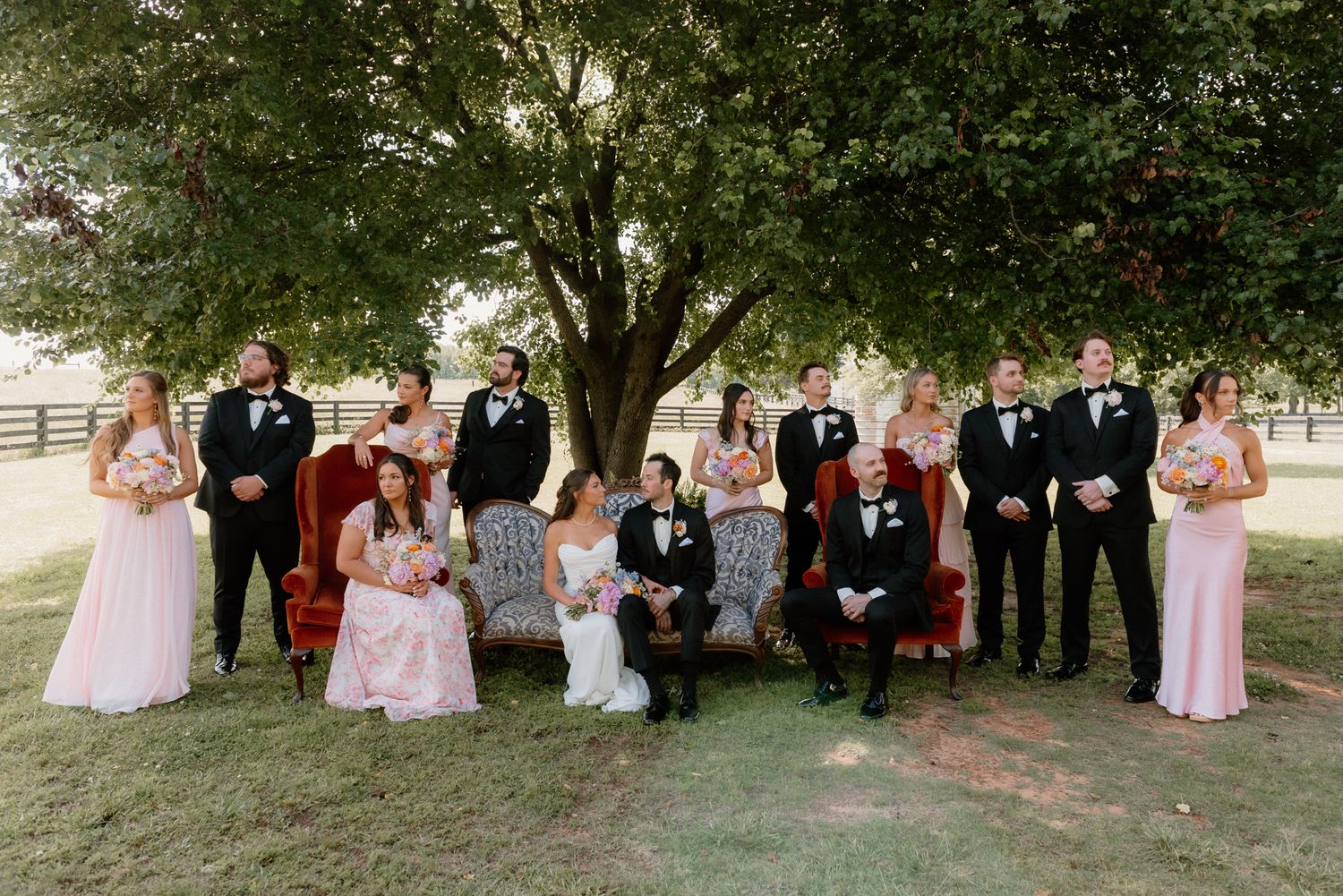 Wedding party in pink bridesmaid dresses and black tuxedos poses outdoors under green trees during summer celebration.